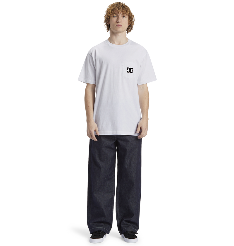 A person with curly hair wears the Men's DC Star Pocket Tee by DC Shoes—a relaxed fit white cotton skate shirt with a small logo—paired with wide dark jeans and black shoes, standing before a plain white background.