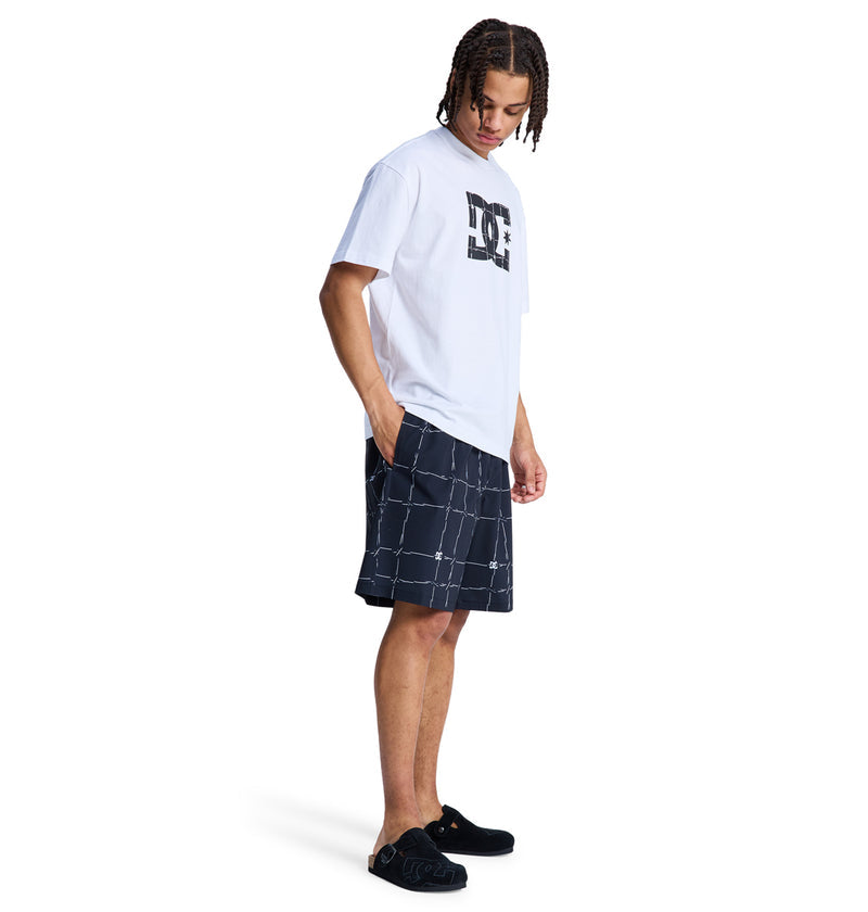 A man stands in profile wearing a white t-shirt, Reebok DC Wired Volley 19 black and white grid-pattern shorts, and black slip-on shoes. He looks down with hands in his pockets against a plain white background.