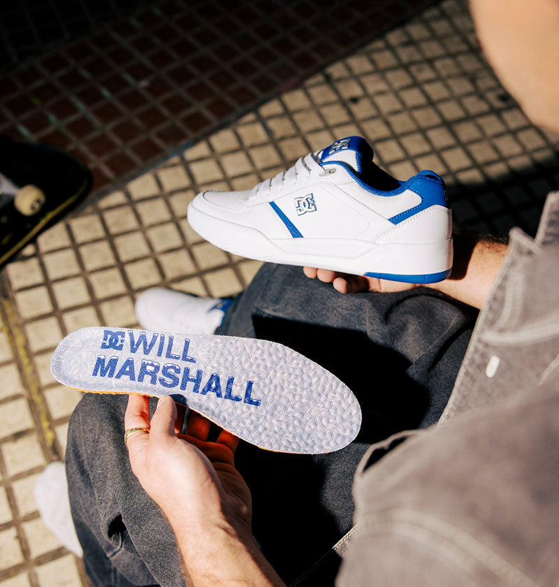 A person holds a pair of DC Shoes Men's Fractal S Sneakers in white and blue, along with the insole featuring pro skater Will Marshall's name printed in bold blue letters. Dressed in dark pants and seated on patterned pavement, they highlight the sneakers' street-ready style.