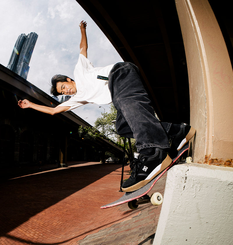 Wearing DC Shoes Men's Fractal S Shoes, a skateboarder in a white shirt and black pants highlights street-ready style while performing a trick on the edge of a concrete ledge, one hand raised with a city building in the background.
