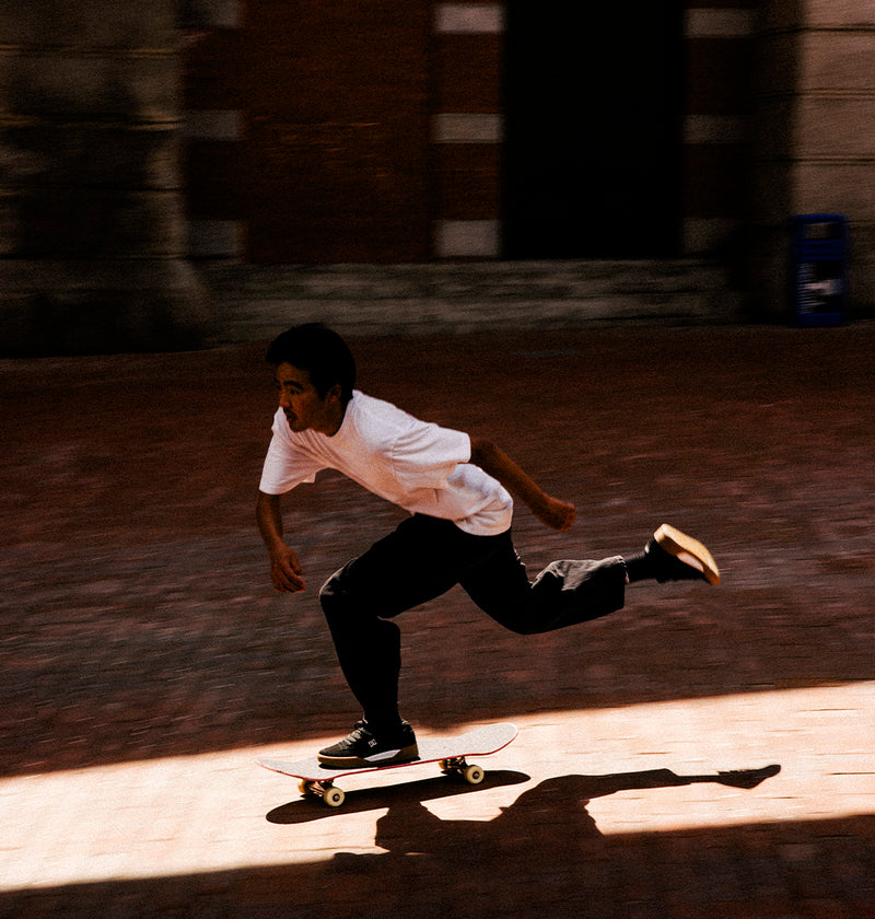 Wearing DC Shoes' Men's Fractal S Shoes, a skateboarder swiftly glides across a sunlit brick pavement, their clear shadow trailing behind—a perfect display of street-ready style and performance-driven comfort.