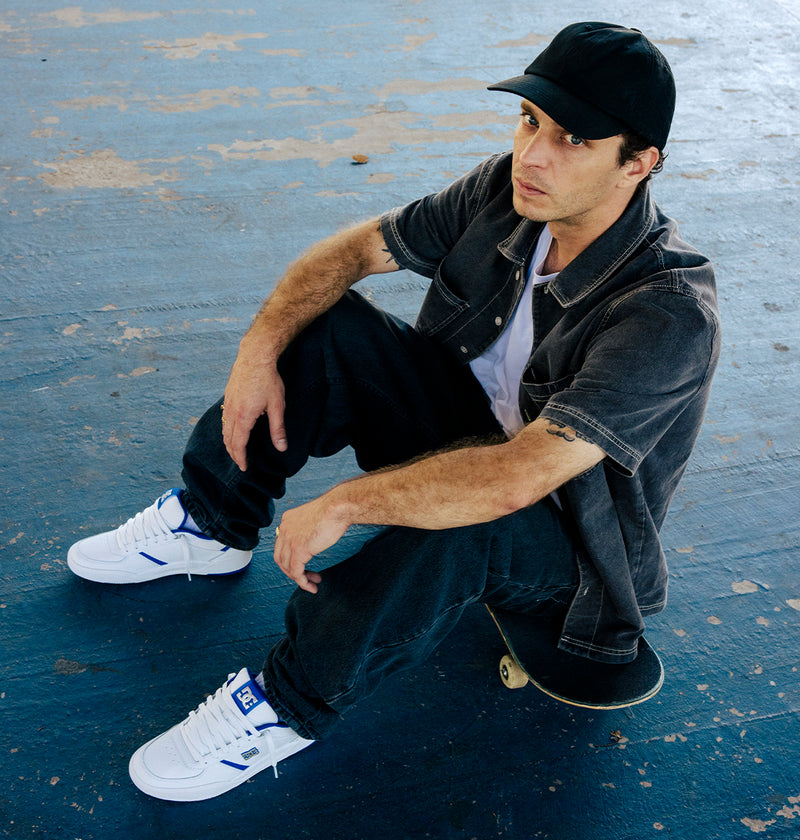 A man wearing a black cap, denim shirt, and DC Shoes Men's Fractal S Shoes sits on a skateboard atop a blue concrete surface, looking up at the camera and channeling street-ready style inspired by pro skater Will Marshall.