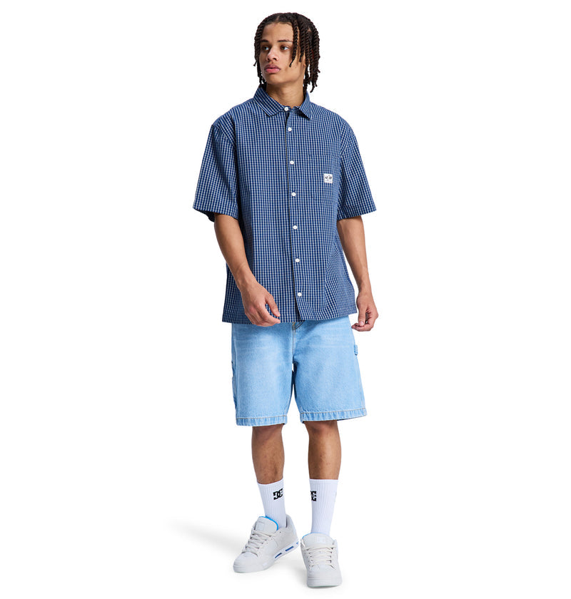 A person wearing the Reebok Game On Ss short-sleeve blue checkered button-up shirt, light blue denim shorts, white crew socks, and white sneakers stands against a plain white background.