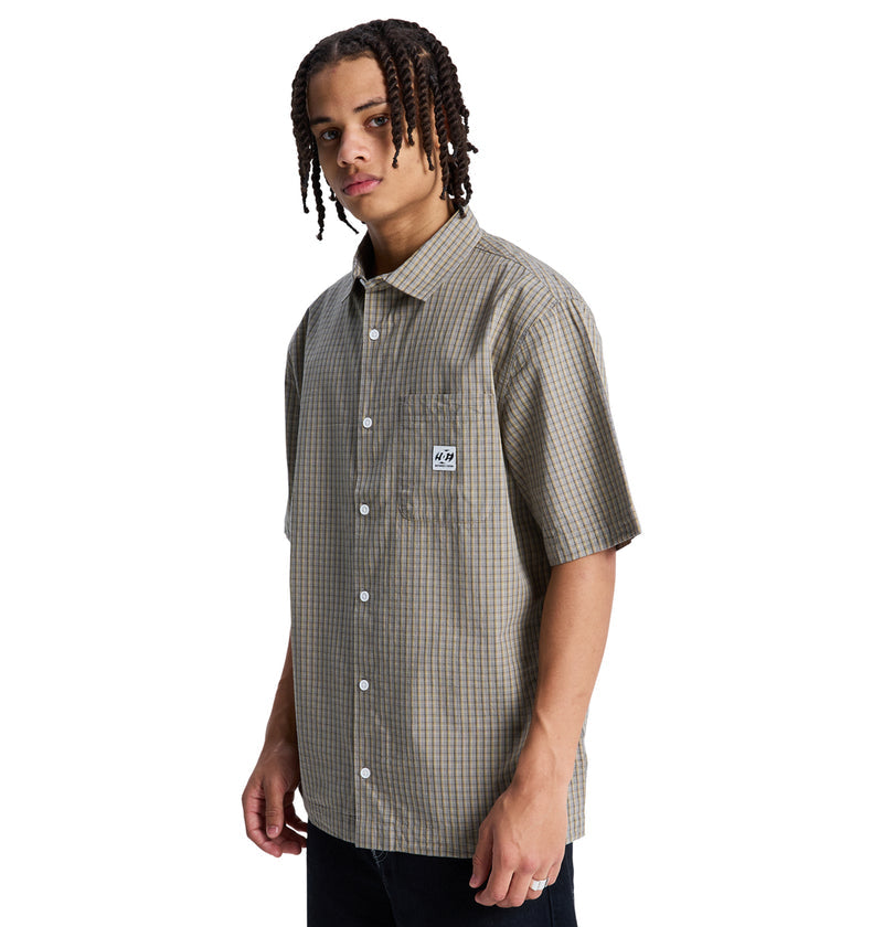 A person with braided hair wears the Reebok Game On Ss short-sleeve, button-up plaid shirt featuring a small patch on the chest pocket, standing against a plain white background.