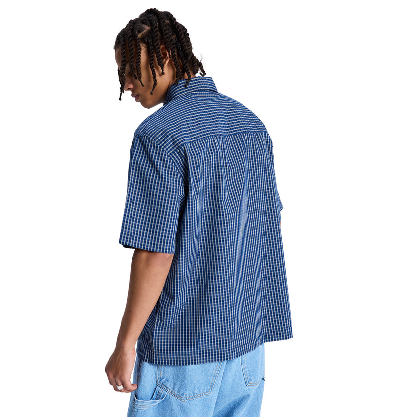 A person with short, twisted hair is seen from behind in a blue and white checkered Reebok Game On Ss short-sleeve shirt and light blue jeans, standing against a plain white background.