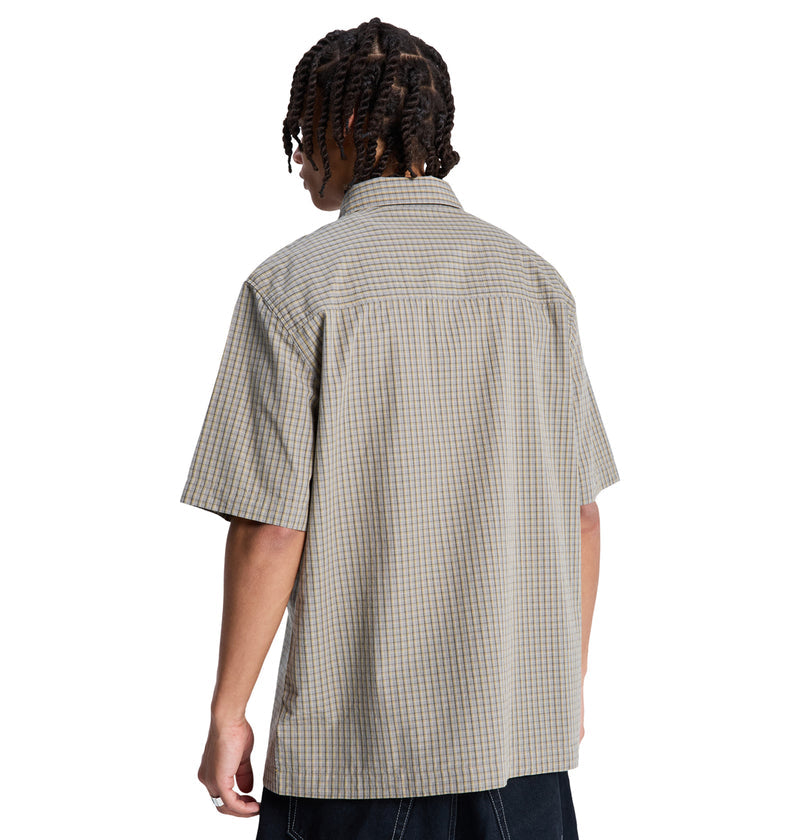 A person with short, twisted hair is seen from behind wearing the Reebok Game On Ss, a short-sleeve loose-fit beige plaid shirt, paired with dark pants against a white background.
