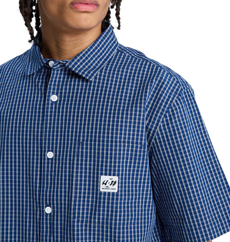 A person wearing the Reebok Game On Ss blue and white checkered short-sleeve button-up shirt with a white chest pocket label. Their face is partially visible.
