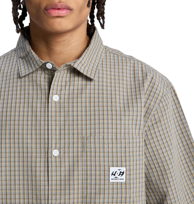 A person wears a beige and blue plaid Reebok Game On Ss button-up shirt with a chest pocket and rectangular logo patch. Only their lower face and upper torso are visible.
