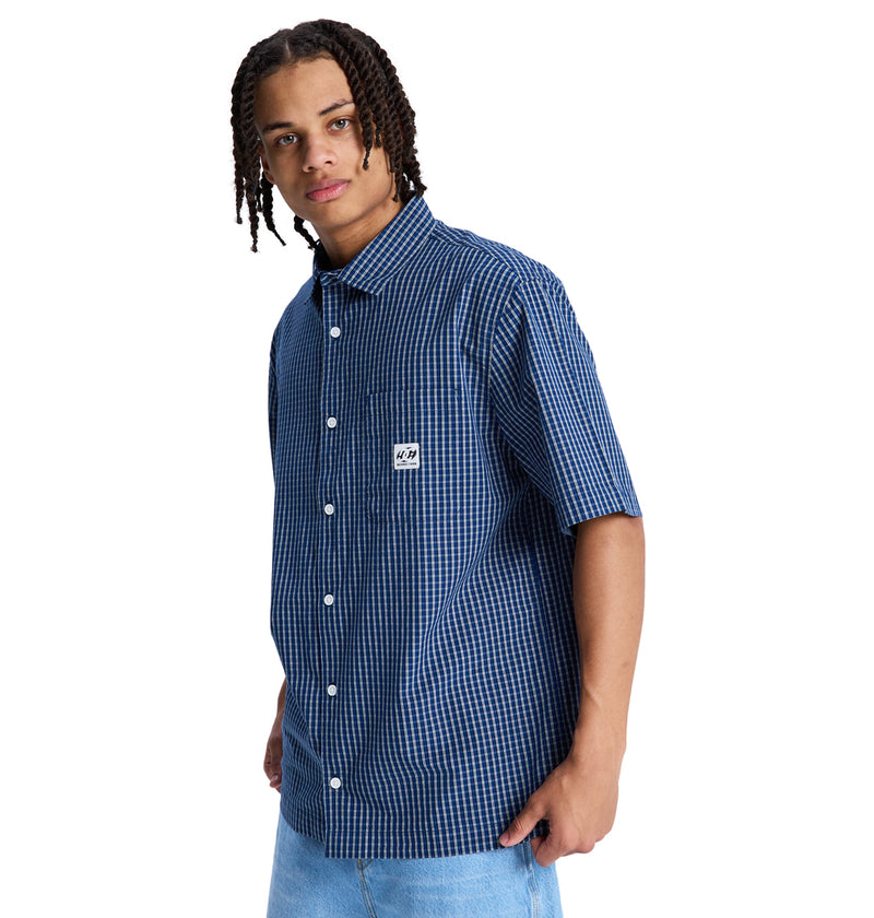 A person with medium-length braided hair wears the Reebok Game On Ss blue and white checkered short-sleeve button-up shirt and light blue jeans, standing against a plain white background.