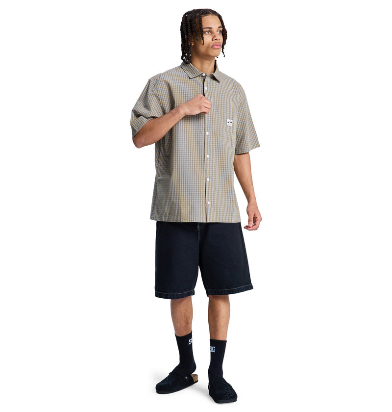 A person with braided hair models the Reebok Game On Ss short-sleeved, checked button-up shirt, black knee-length shorts, black socks, and sandals against a plain white background.