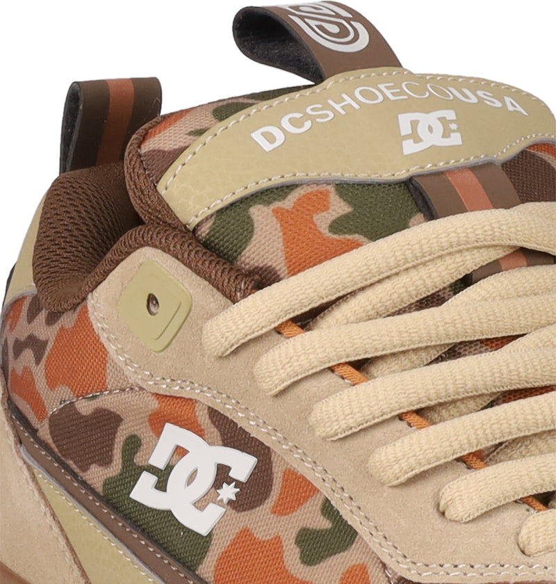 Close-up of the Men's JS 2 Shoes by DC Shoes, inspired by DC heritage and John Shanahan. Features a tan suede upper, orange-green camo pattern, beige laces, white DC logo on the side, and a DCSHOECOUSA tongue label.