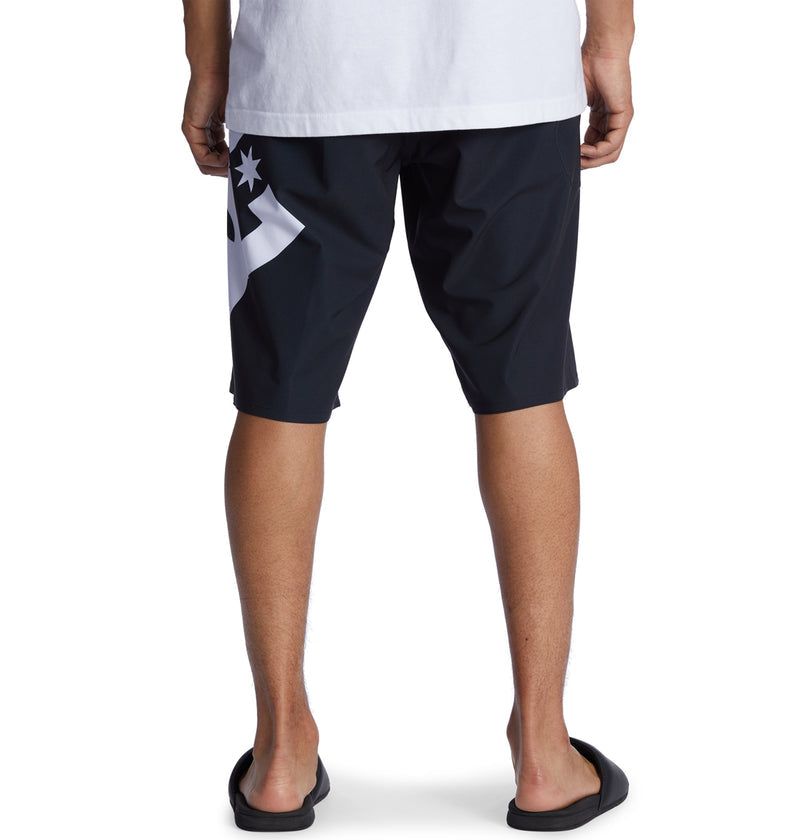 A person wearing Reebok Lanai 21 black knee-length shorts with a white graphic on the back, a white T-shirt, and black slide sandals, stands with their back to the camera.