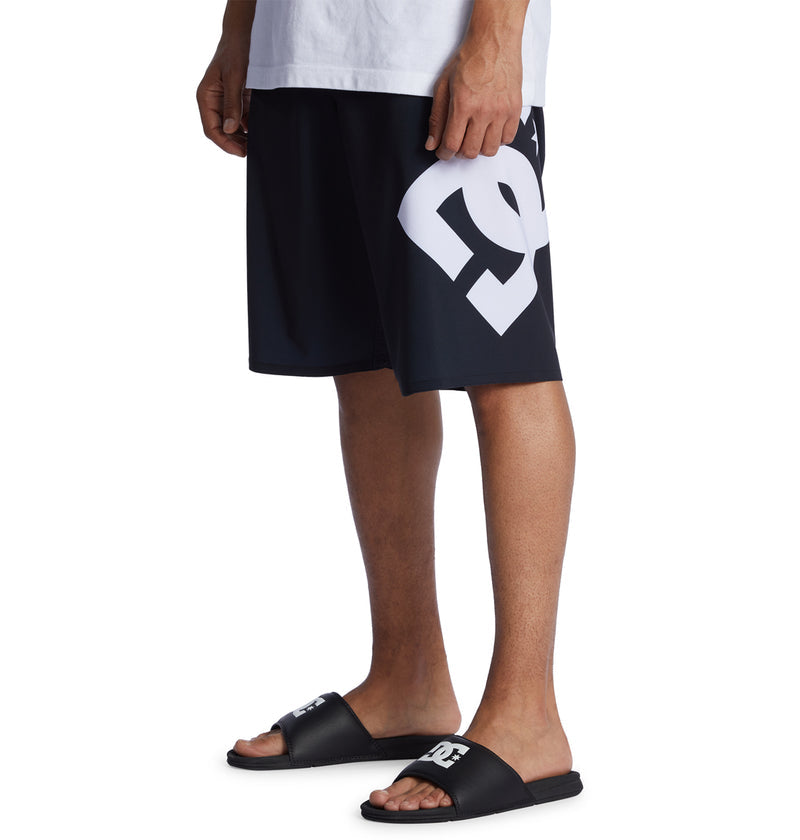 A person wearing black Reebok Lanai 21 shorts with a large white logo, a white shirt, and black slide sandals stands on a white background.