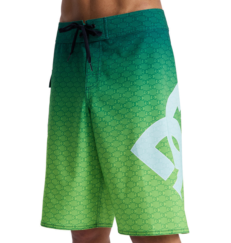 A person wearing Reebok Lanai 21 green gradient board shorts with a black drawstring, a large white stylized DC logo on the left leg, subtle repeating pattern, and a length just above the knees.