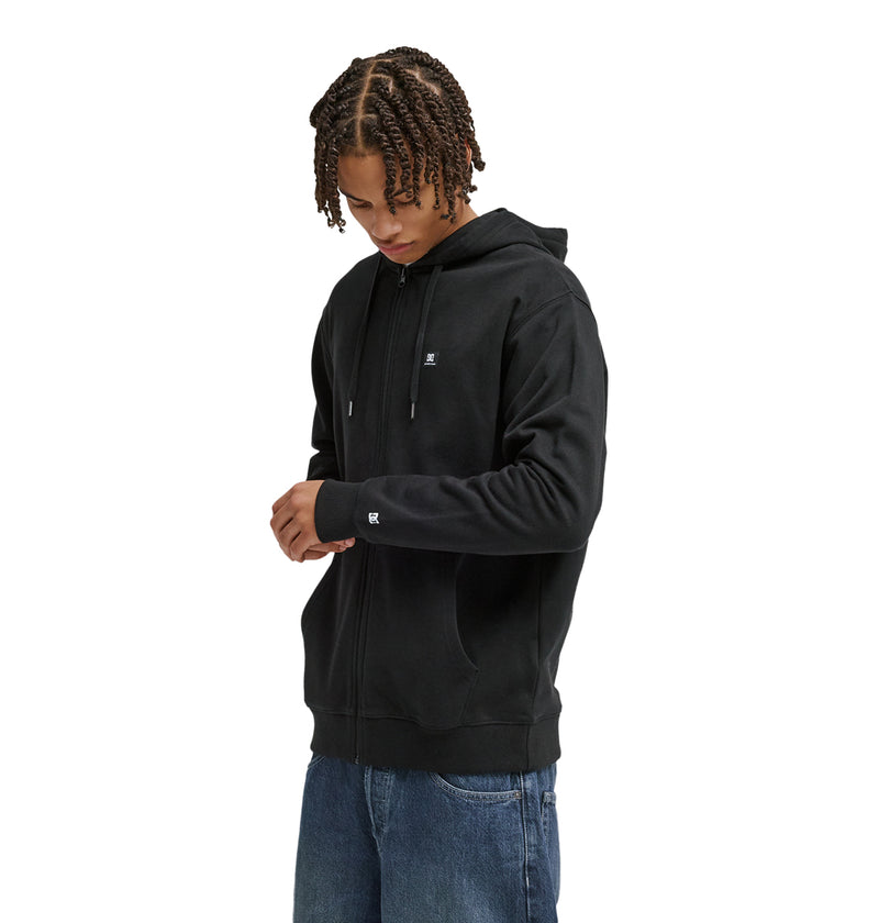 A person with short, braided hair wears the Reebok Lorion Zh black zip-up hoodie and blue jeans, looking down and adjusting their sleeve against a plain white background.