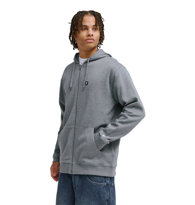 A young person with curly hair models the Reebok Lorion Zh gray zip-up hoodie and blue jeans, standing with one hand in their pocket and looking to the side against a plain white background.