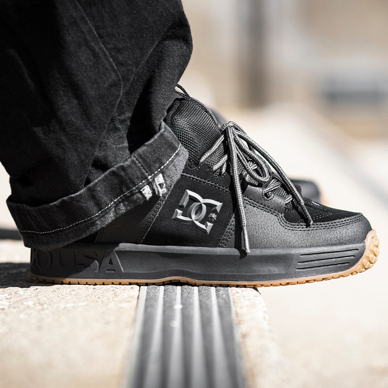 Close-up of a person wearing Men's Lynx OG Shoes by DC Shoes with light brown soles, featuring a bold DC logo on the side. Perfect for skateboarding, the shoes are paired with black pants and stand out on a concrete surface.