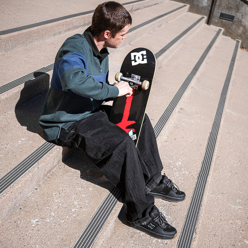 A person sits on outdoor concrete stairs, adjusting a skateboard wheel. They're wearing a green and blue sweater, black pants, and DC Shoes Men's Lynx OG Shoes as sunlight casts shadows over the steps.