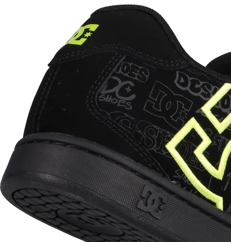 Close-up of the heel of a black DC Shoes Men's Net Shoe with neon yellow accents and a grey logo. This sneaker features strong DC branding, durable fabrics, and a textured black sole with a small DC Shoes logo on the side.