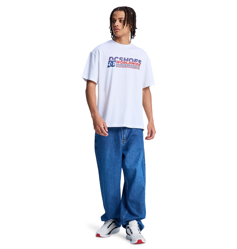 A young man with short dreadlocks wears the Reebok Queltra Lhss white graphic T-shirt, loose blue jeans, and white sneakers with red and gray details, standing against a plain white background.