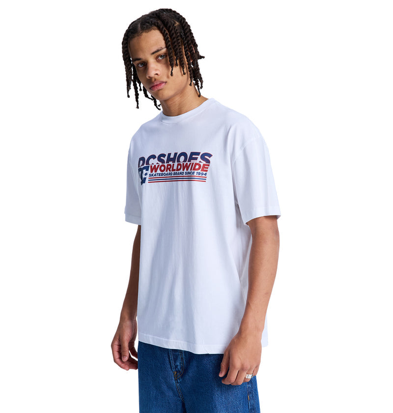 A person with braided hair wears the Reebok Queltra Lhss white t-shirt with a red and blue graphic on the chest, paired with blue jeans, standing against a plain white background.