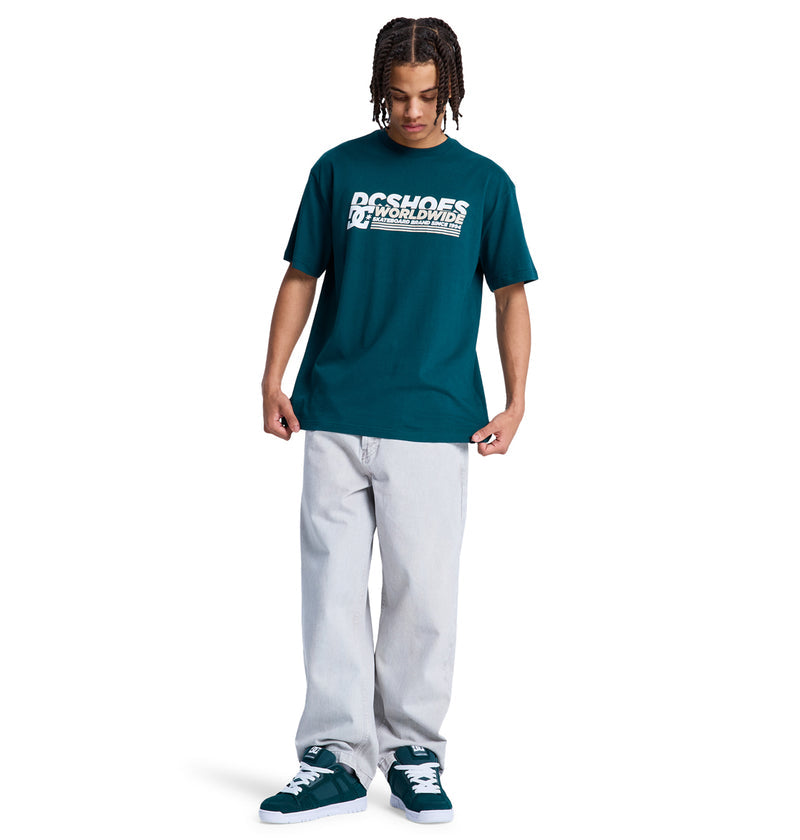 A young man wearing a teal Reebok Queltra Lhss t-shirt, light gray baggy jeans, and teal sneakers stands against a white background, looking down with his hands at his sides.