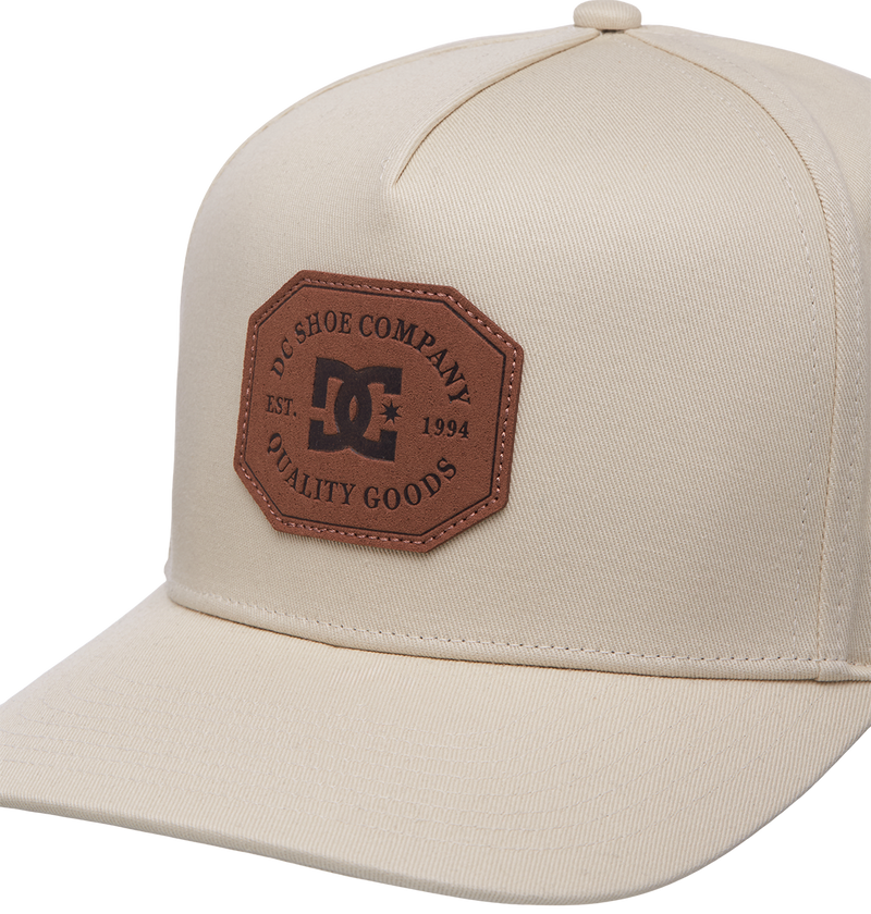 The Men's Reynotts Cap by DC Shoes is a beige skate cap featuring a brown hexagonal patch on the front with DC branding, reading "DC Shoe Company Quality Goods Est. 1994," and has a classic curved brim.