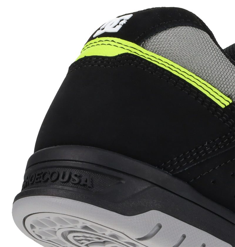 Close-up of the heel of DC Shoes Men's Stag Shoes with a dynamic black upper, gray mesh panel, neon yellow stripe, and white DC logo. The durable gray cupsole has textured details and SHOECOUSA text on the back.