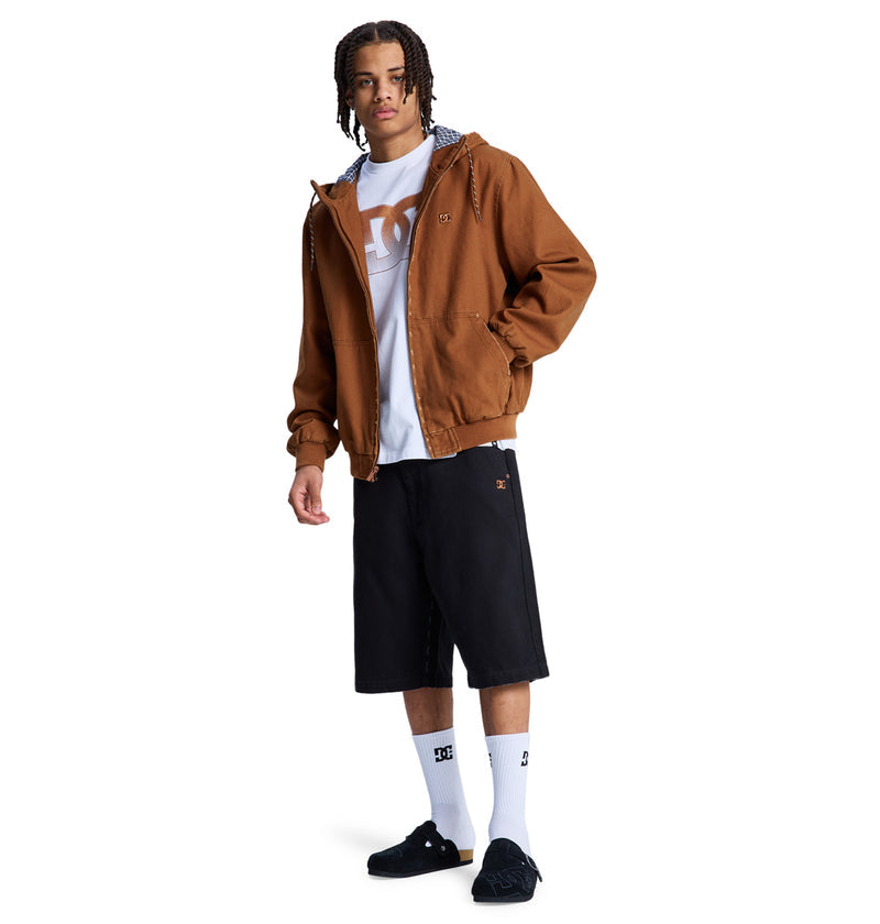 A person wears the Reebok Stressed 5 Pocket brown jacket over a white T-shirt, black knee-length shorts, DC logo white socks, and black sneakers while standing against a white background.