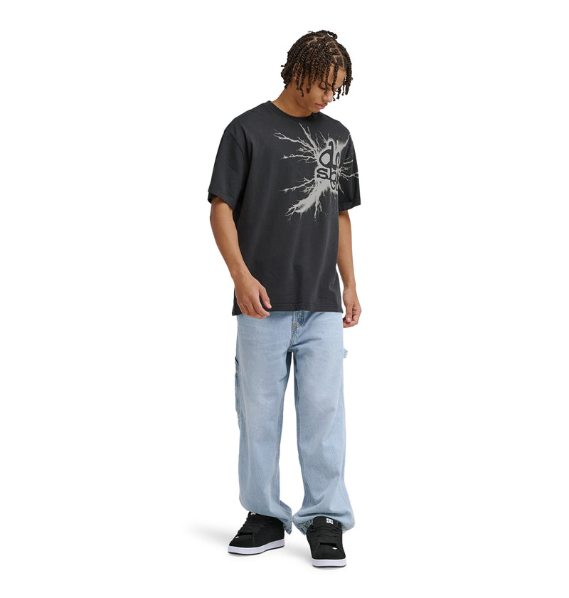 A person with short braids wearing a black graphic t-shirt, light blue baggy jeans, and Reebok Strike Twice Hss sneakers stands looking down against a plain white background.