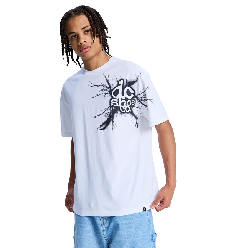 A person with braided hair wears a loose white Reebok Strike Twice Hss t-shirt with a black graphic logo and blue denim shorts, standing against a white background.