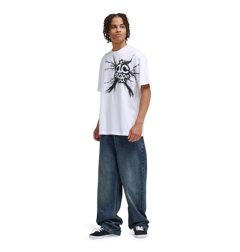 A young man with short dreadlocks wears the Reebok Strike Twice Hss graphic T-shirt, loose blue jeans, and black sneakers, standing against a plain white background.