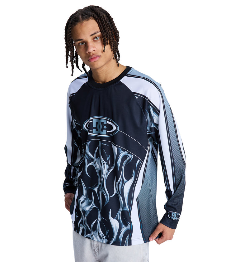 A young person with braided hair models the Reebok Sylem 2 Ls, a long-sleeve black and white graphic shirt with a bold flame design, standing against a white background.