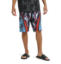 A person wearing Reebok Sylem 21 black slide sandals and bold flame-patterned knee-length shorts, with only their legs and lower torso visible, stands against a white background.