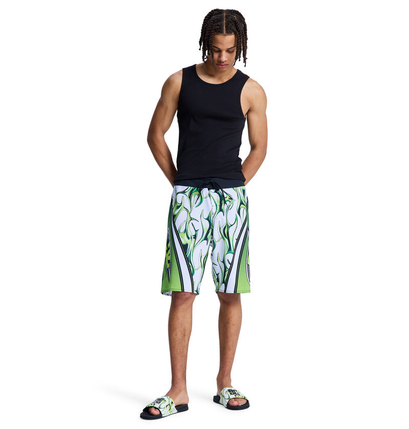 A person with medium skin and braided hair wears the Reebok Sylem 21 black tank top, green and white graphic shorts, and matching slide sandals, standing with hands in pockets while looking down against a white background.